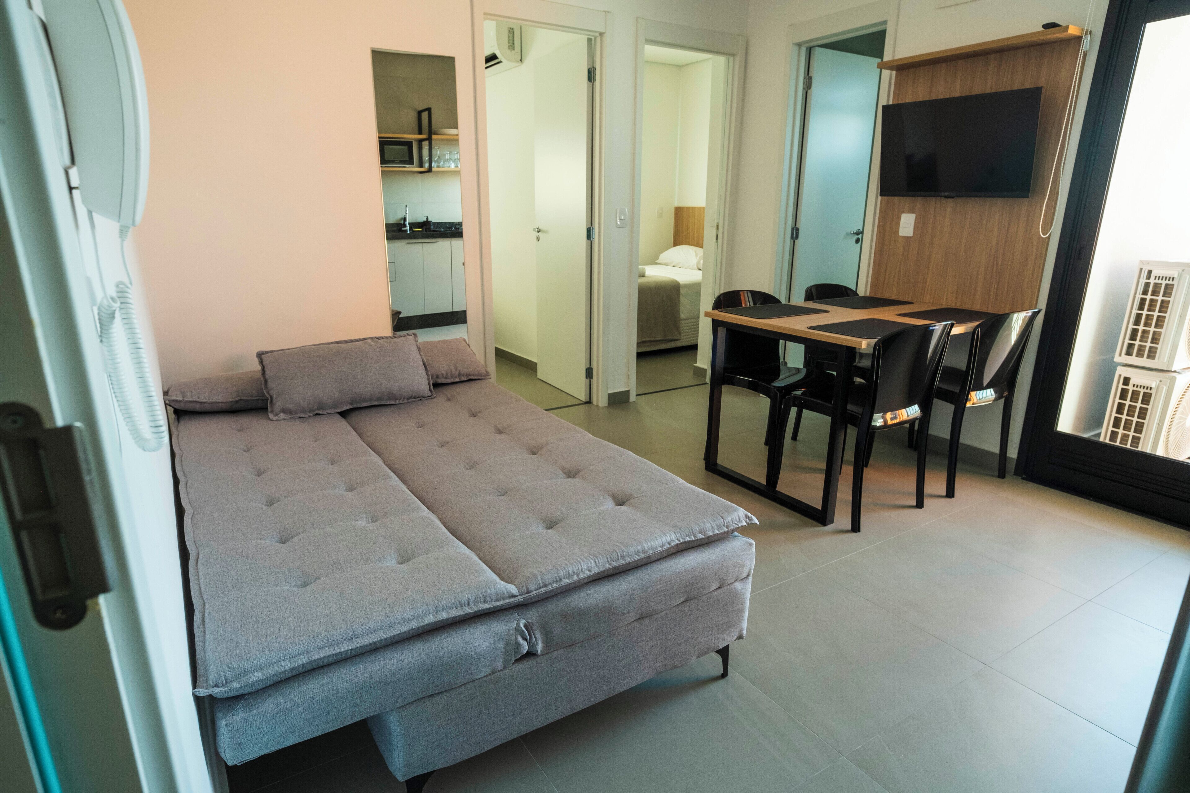 Habitación pequeña con cama, escritorio y silla. Hay un televisor en la pared y una puerta que da a otra habitación.