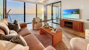 Ein geräumiges Wohnzimmer mit großem Fenster mit Meerblick, einer bequemen Couch, einem Couchtisch mit Dekoration und einem an der Wand befestigten Flachbildfernseher.