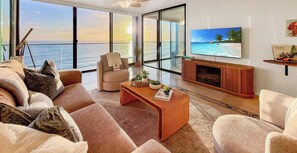 Ein geräumiges Wohnzimmer mit großem Fenster mit Meerblick, einer bequemen Couch, einem Couchtisch mit Dekoration und einem an der Wand befestigten Flachbildfernseher.