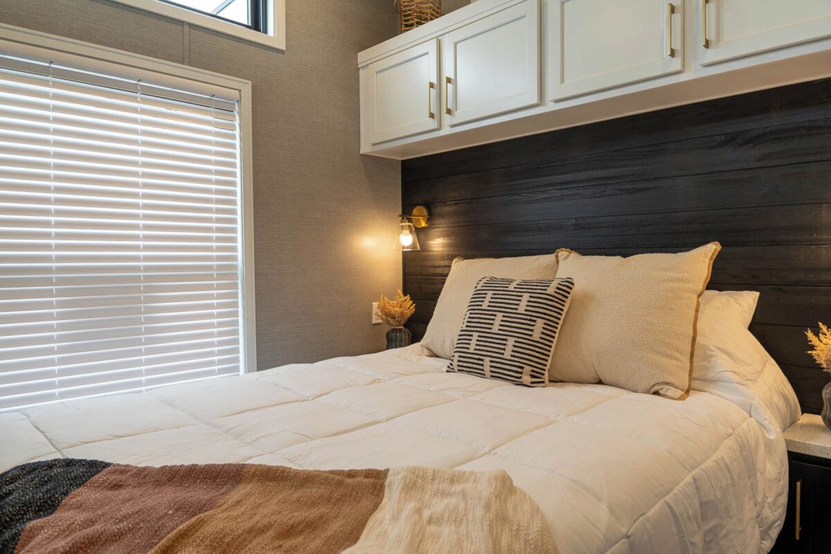A bedroom with a bed, a window with blinds, a nightstand with a lamp, and a headboard with cabinets.