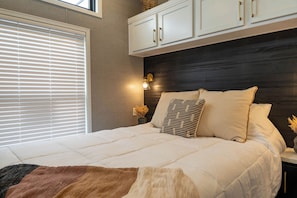 Un dormitorio con cama, ventana con persianas, mesita de noche con lámpara y cabecera con armarios.