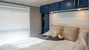A bedroom with a bed, pillows, a window with blinds, and blue cabinets above the bed. - Frieden Saugatuck (Saugatuck)