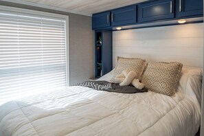 A bedroom with a bed, pillows, a window with blinds, and blue cabinets above the bed. - Frieden Saugatuck (Saugatuck)