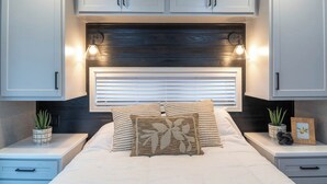 A bedroom with a bed, two bedside tables, a headboard with built-in lighting, and a window with blinds. - Frieden Saugatuck (Saugatuck)
