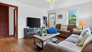 A living room with a television, a coffee table with decorative items, a sofa, and a window with a view of greenery. - Creekside Cabin by Avantstay Creek Swims + Trout Fishing Firepit Deck Views Near Hunter Mtn (Jewett)