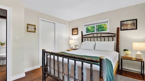 A bedroom with a wooden bed frame, a window with a view of greenery, a bedside table with a lamp, and a door. - Creekside Cabin by Avantstay Creek Swims + Trout Fishing Firepit Deck Views Near Hunter Mtn (Jewett)