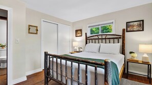 A bedroom with a wooden bed frame, a window with a view of greenery, a bedside table with a lamp, and a door. - Creekside Cabin by Avantstay Creek Swims + Trout Fishing Firepit Deck Views Near Hunter Mtn (Jewett)