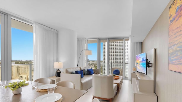 Ein modernes Hotelzimmer mit Essbereich, einer Sofagruppe, einem Fernseher und großen Fenstern mit Blick auf die Stadt.