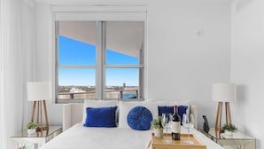 Ein Hotelzimmer mit einem großen Fenster, einem Bett mit blauen Kissen, einem Nachttisch mit einer Flasche und Gläsern sowie einer kleinen Pflanze.