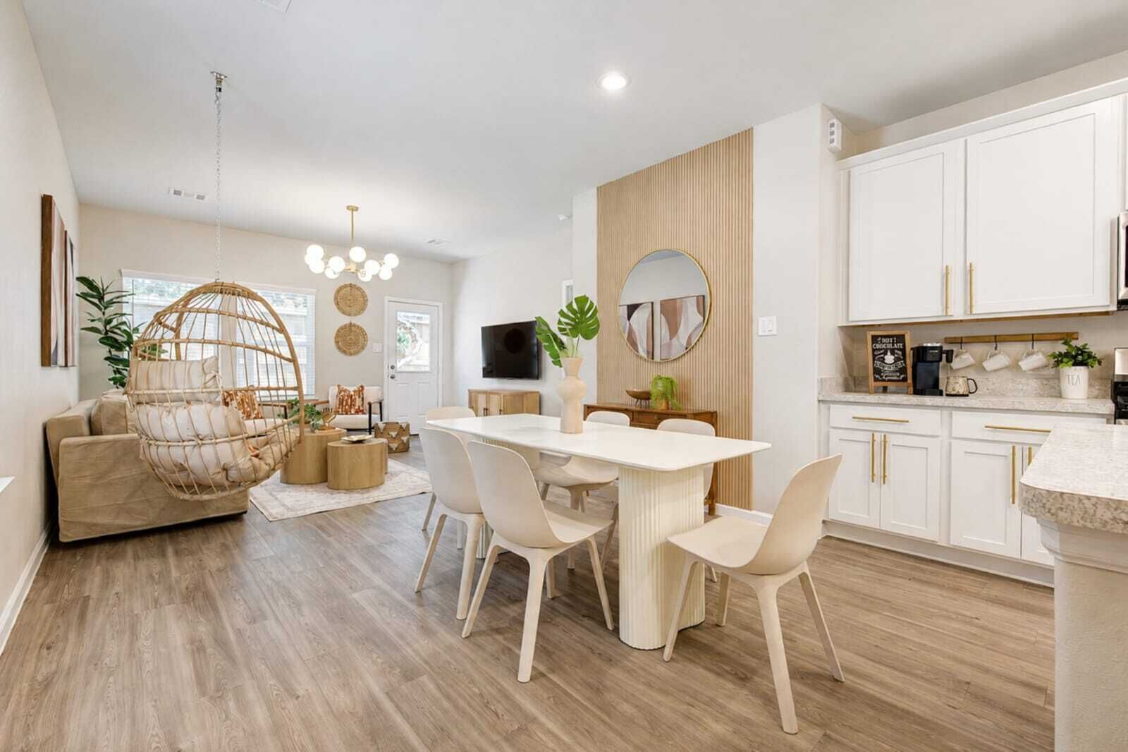 Een moderne keuken met witte kasten, een eethoek met een witte tafel en stoelen, en een hanglamp.
