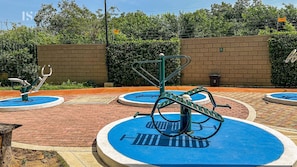 Ein Trainingsbereich im Freien mit verschiedenen Fitnessgeräten, darunter eine Beinpresse und ein Handbike, umgeben von einer Backsteinmauer und Pflanzen.
