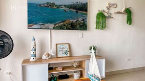 A room with a television displaying a coastal scene, a small shelf with decorative items, and a wall-mounted fan. - Comfortable Apartment 2Bed/2Bath - Bello Horizonte - Santa Marta (Santa Marta)