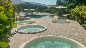 A pool area with multiple hot tubs and lounge chairs, surrounded by greenery and a mountainous backdrop.
