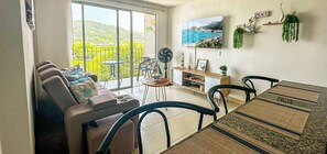 A living room with a sofa, chairs, a dining table, and a view of a green landscape through large windows. - Comfortable Apartment 2Bed/2Bath - Bello Horizonte - Santa Marta (Santa Marta)