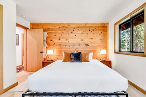 Une chambre à coucher avec une tête de lit en bois, un grand lit avec une literie blanche, deux lampes de chevet et une fenêtre donnant sur des arbres.
