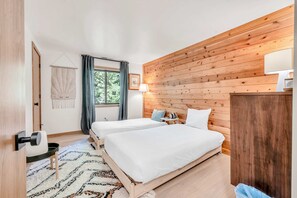Ein Zimmer mit zwei Betten, einer Holzwand, einem Fenster mit Vorhängen, einem kleinen Tisch und einer Wandlampe.