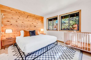 Ein Schlafzimmer mit einer Wand aus Holzlatten, einem Bett mit weißer Bettwäsche, zwei Nachttischlampen, einem Babybett und einem Fenster mit Blick auf Bäume.