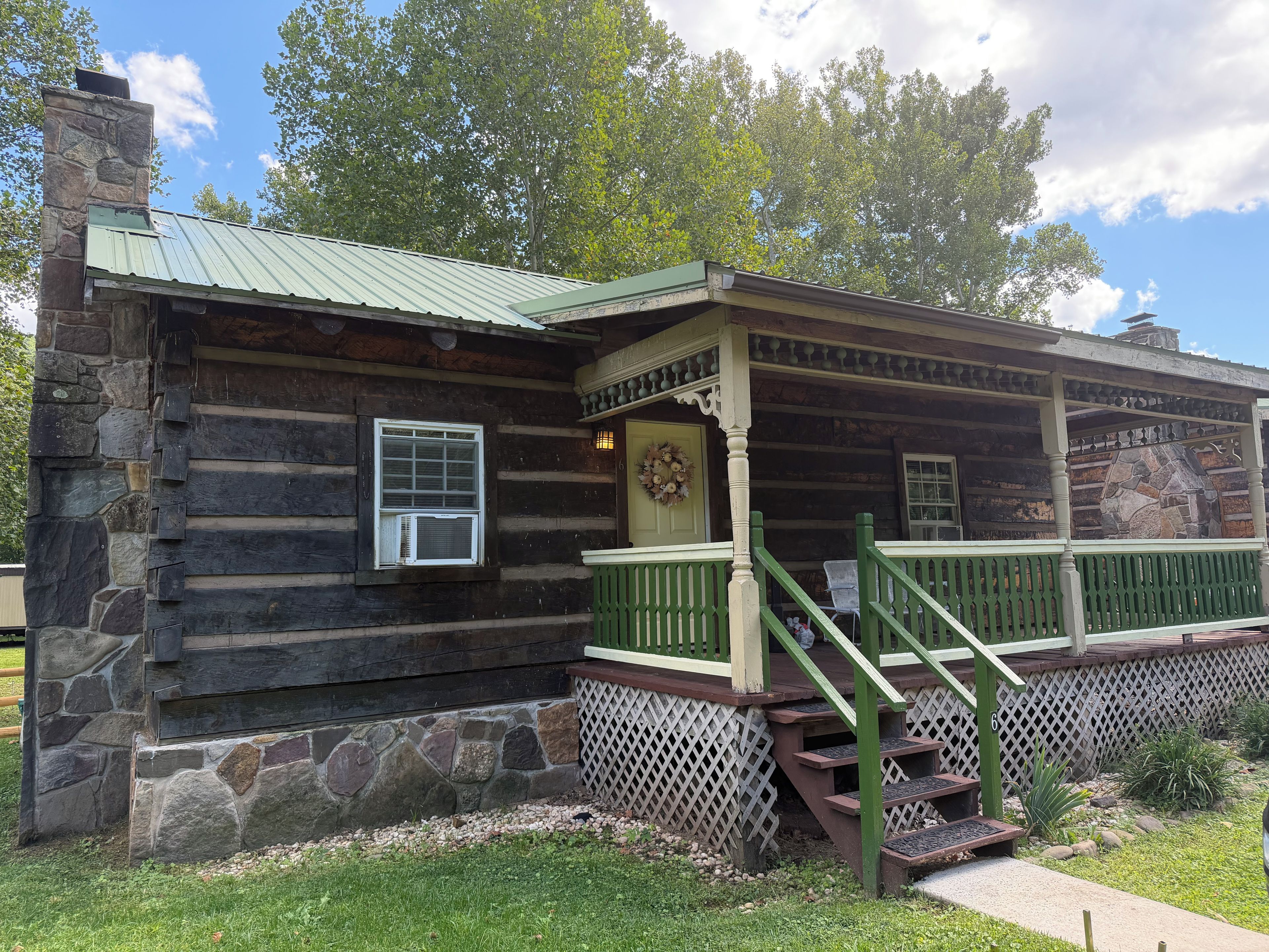 A log cabin with a stone foundation, a porch with green railings, and a chimney.