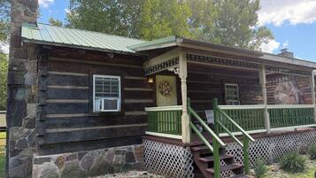 A log cabin with a stone foundation, a porch with green railings, and a chimney.