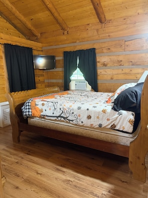 A wooden cabin room with a bed, a television, and a window with curtains.