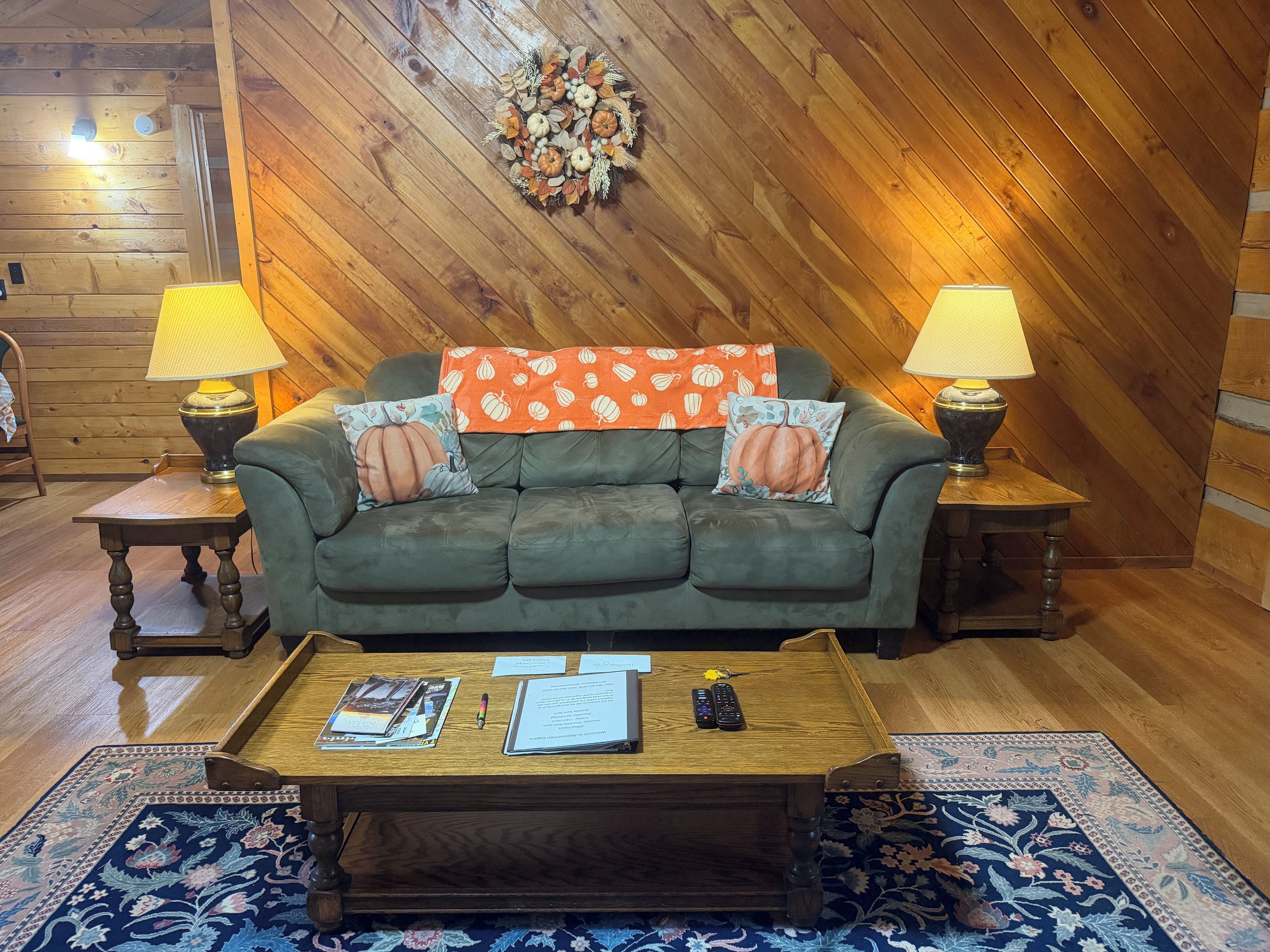 A cozy living room with a wooden ceiling, a green sofa, two side tables with lamps, a coffee table with magazines and remotes, and a decorative wall hanging.