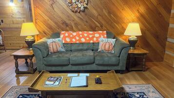 A cozy living room with a wooden ceiling, a green sofa, two side tables with lamps, a coffee table with magazines and remotes, and a decorative wall hanging.