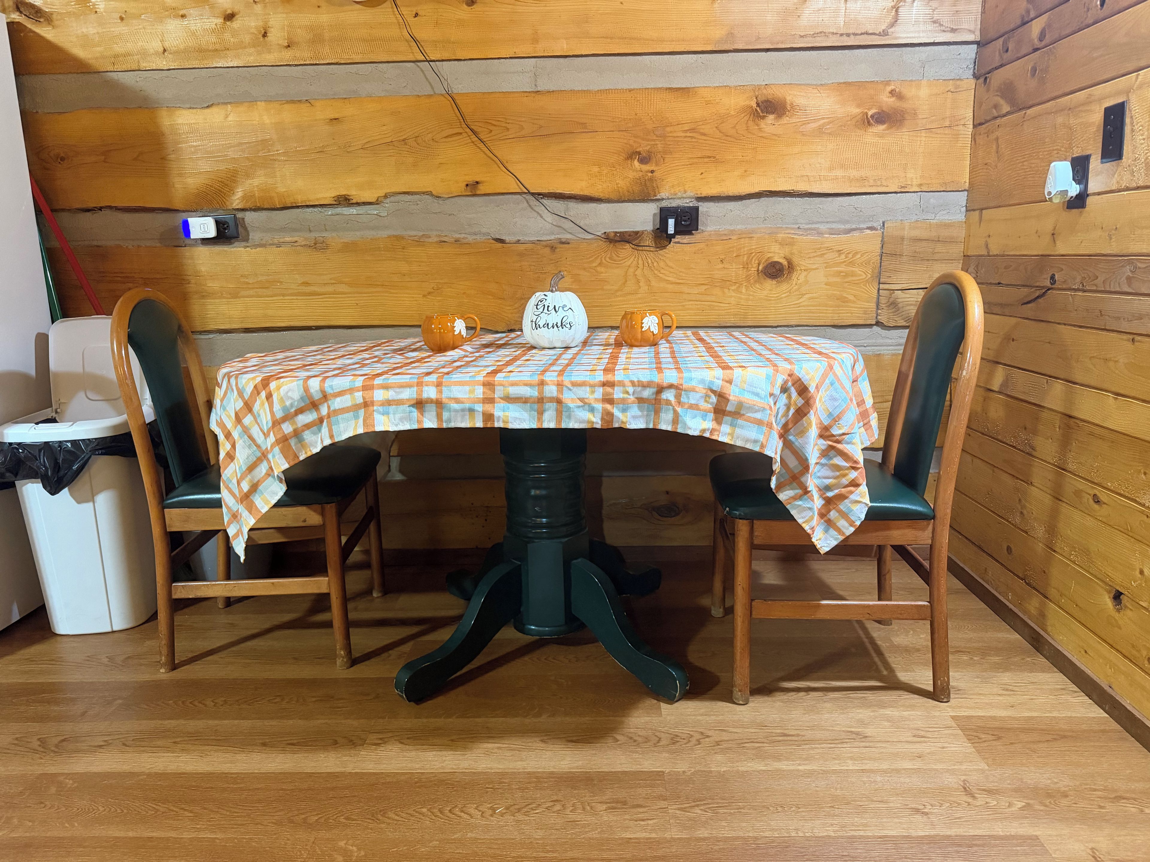 A wooden dining table with a plaid tablecloth, two chairs, a water jug, and a sign that says 'Give thanks.'