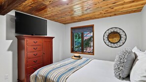 Un dormitorio con techo de madera, un televisor sobre un mueble, una ventana con vistas y un espejo decorativo.