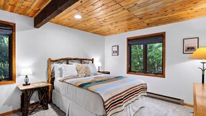 Een slaapkamer met een houten plafond, een bed met een gestreept dekbed, twee nachtlampjes en een raam met uitzicht op bomen.