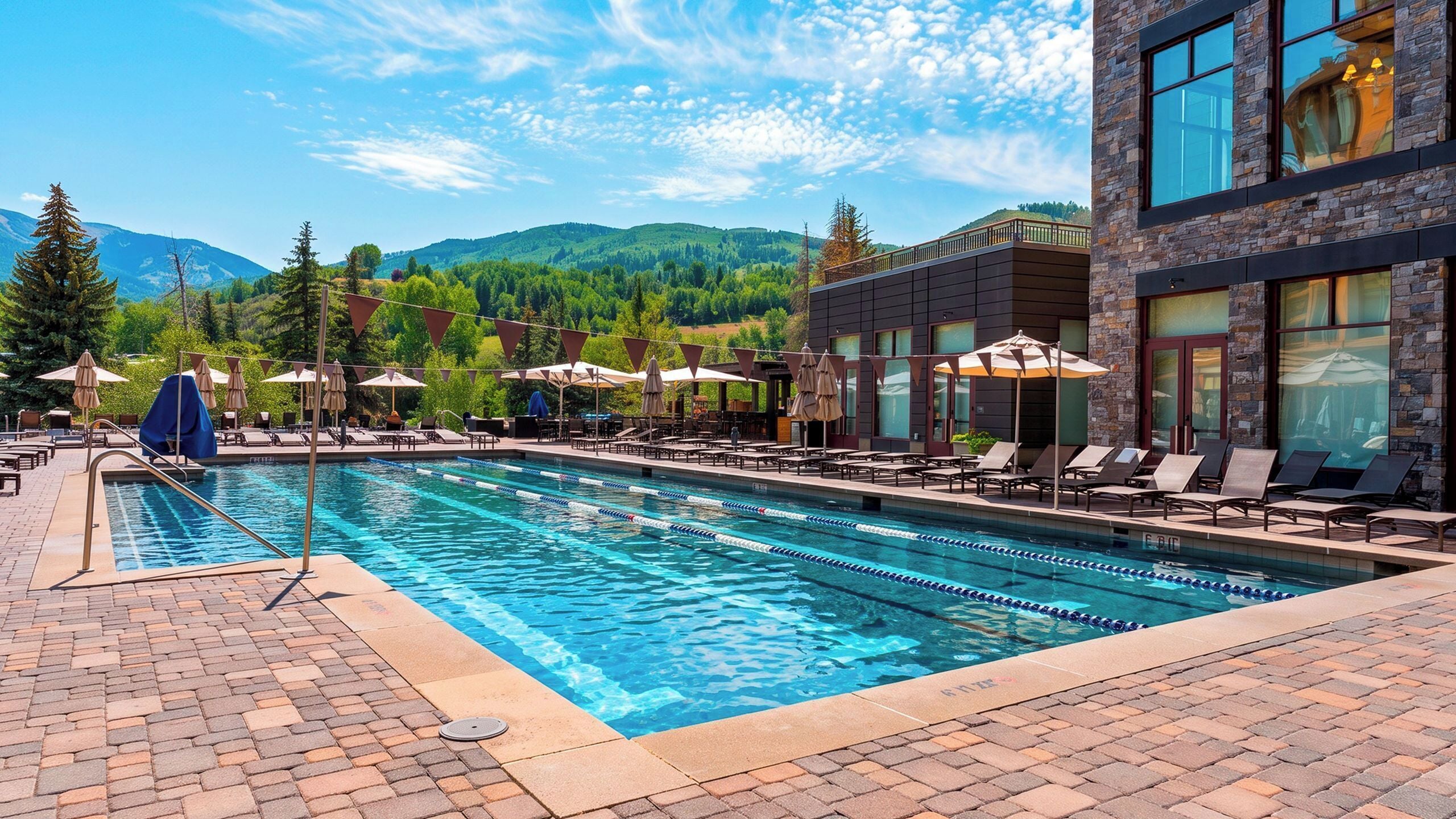 A swimming pool with lounge chairs and umbrellas, surrounded by a brick patio and a building with large windows.
