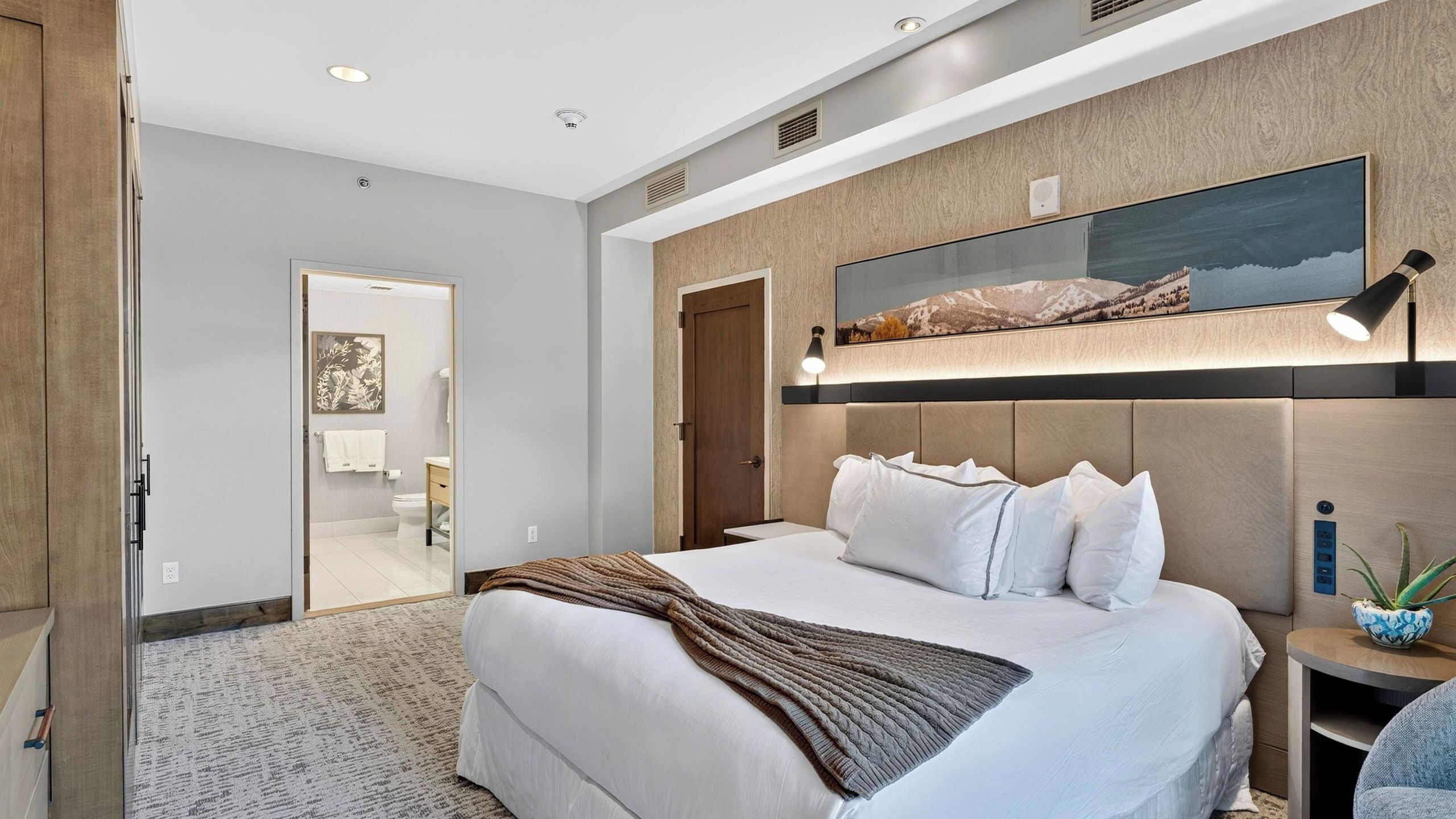 Habitación de hotel con una cama, una mesita de noche con lámpara, televisor montado en la pared y una puerta que da al baño.