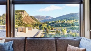 A room with a large window offering a view of a mountainous landscape, a sofa with multiple cushions, and a door with a chain. - Riverfront Perch by Avantstay Balcony + Resort Pool River + Mtn Views (Avon)