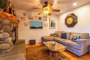 A living room with a stone wall, a ceiling fan, a sofa with decorative pillows, a coffee table, and a wall clock. - Tahoe Village Condo | Walk to Heavenly Stagecoach (Stateline)
