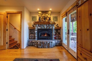 Une cheminée en pierre avec un feu de bois, un plancher en bois, un escalier et une vue sur l’extérieur grâce à une porte vitrée.