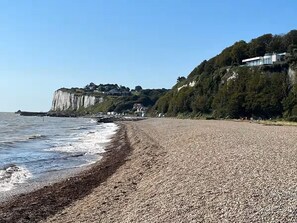 Ein Kiesstrand mit einer Klippe und Häusern im Hintergrund.