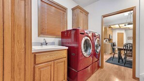 A laundry room with a red washer and dryer, wooden cabinets, a sink, and a window with blinds.