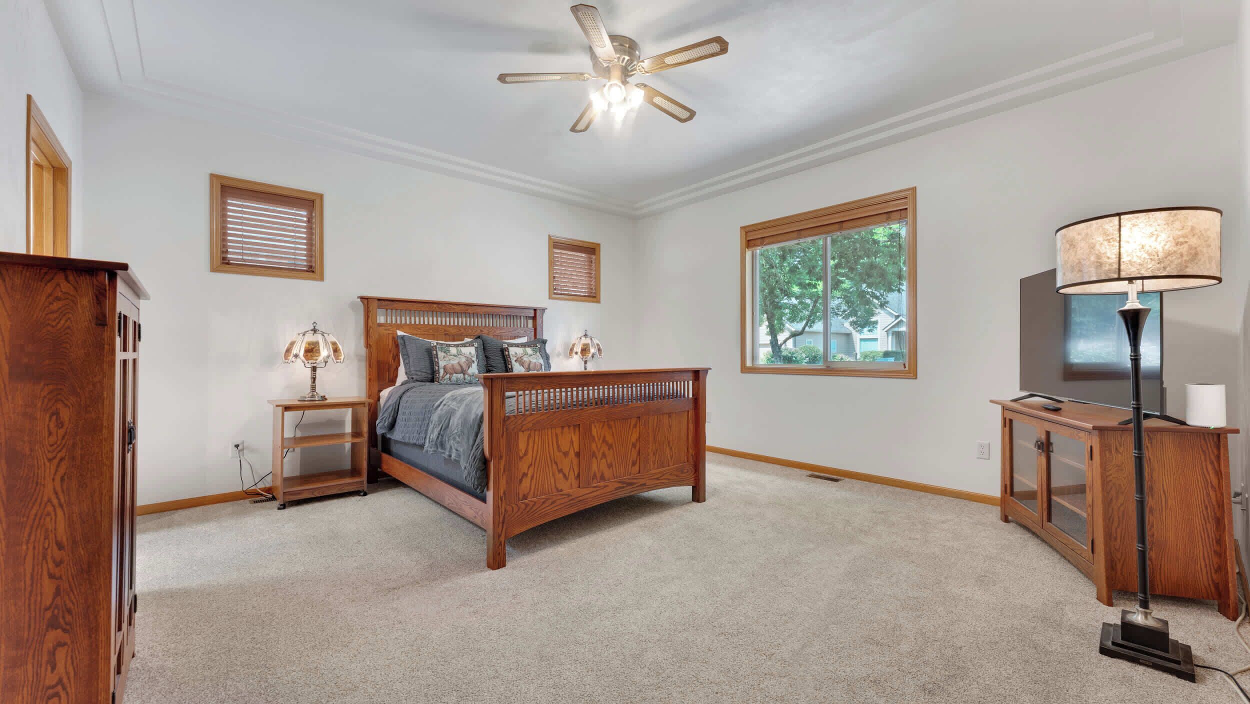 A bedroom with a wooden bed, a ceiling fan, a desk with a lamp, and a window with blinds.