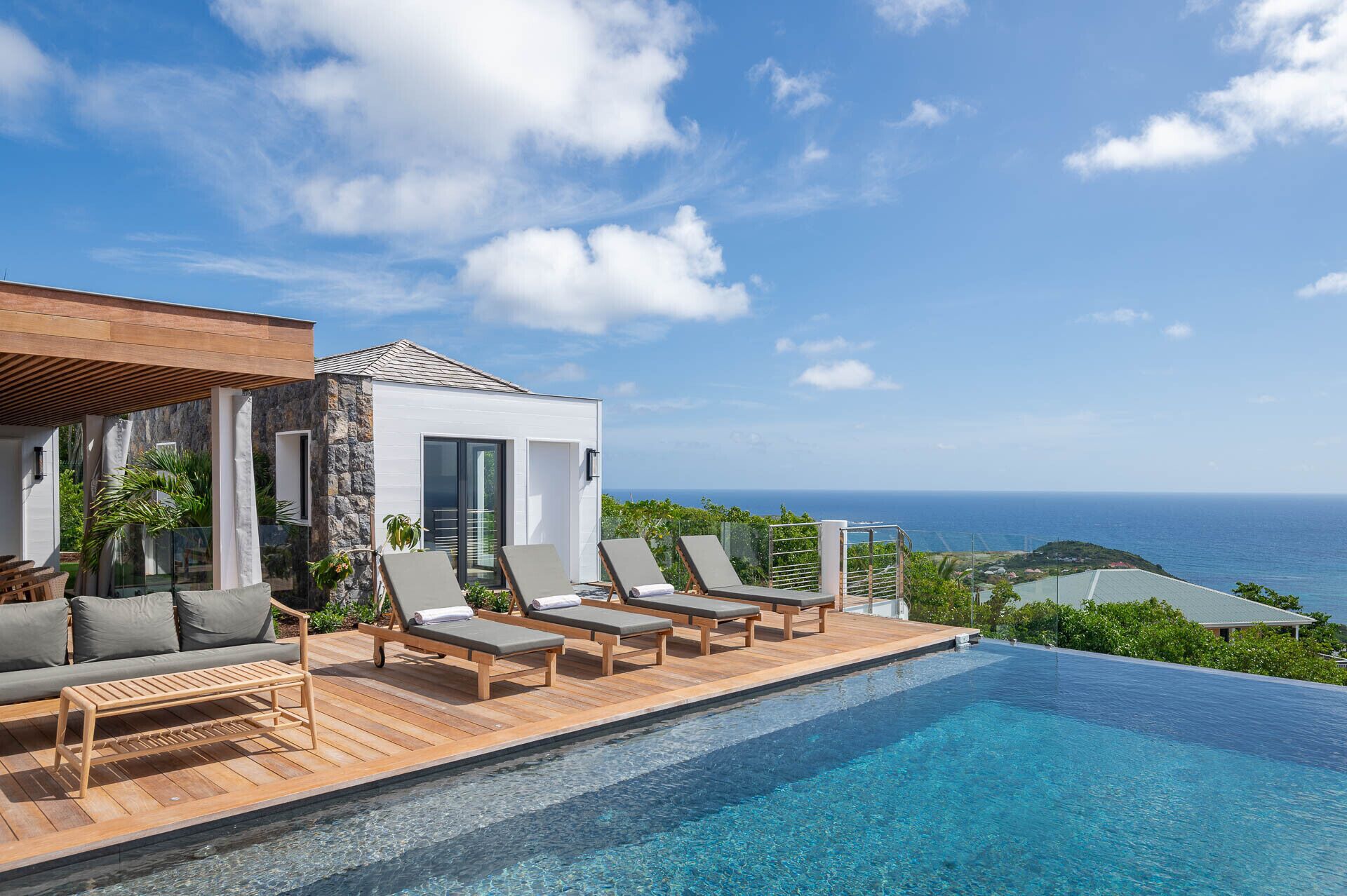 A modern infinity pool with a clear view of the ocean, lounge chairs, and a stone building with a white facade.