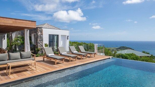 A modern infinity pool with a clear view of the ocean, lounge chairs, and a stone building with a white facade. - Villa Scarlett (4 bedrooms) (Saint Barthélemy)