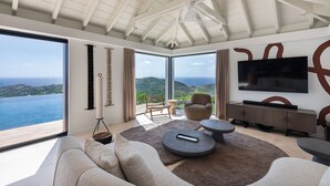 A modern living room with a large window, a flat-screen TV, and a view of the sea. - Villa Scarlett (4 bedrooms) (Saint Barthélemy)
