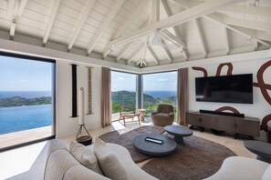 A modern living room with a large window, a flat-screen TV, and a view of the sea. - Villa Scarlett (4 bedrooms) (Saint Barthélemy)