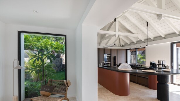 Una cucina moderna con una grande isola, un'area pranzo con una sedia in vimini e vista su un giardino rigoglioso attraverso una porta in vetro.