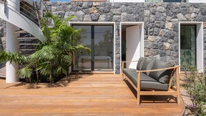 Zona relax esterna moderna con terrazza in legno, parete in pietra e vista su una camera da letto attraverso una porta in vetro.