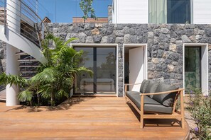 A modern outdoor seating area with a wooden deck, a stone wall, and a view of a bedroom through a glass door. - Villa Scarlett (2 bedrooms) (Saint Barthélemy)