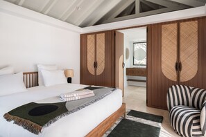 A bedroom with a bed, a bedside lamp, a striped armchair, and a wardrobe with woven doors. - Villa Scarlett (2 bedrooms) (Saint Barthélemy)
