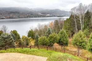 Ein gepflasterter Bereich mit Holzlattenzaun, umgeben von Bäumen und einem See mit Bergen im Hintergrund.