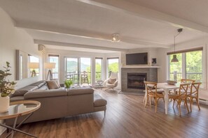 Een ruime woonkamer met een open haard, een bank, een eettafel met stoelen en grote ramen met uitzicht op het landschap.