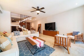 A modern living room with a sofa, coffee table, and chairs, a television, and a staircase leading to an upper level. - Your Home in the Mayan Jungle (Tulum)