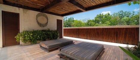 A covered outdoor seating area with two cushioned benches, a wall-mounted decorative item, and a view of greenery.