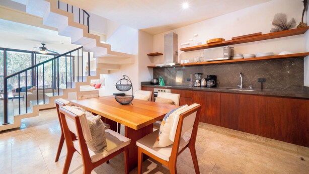 A modern kitchen with a wooden dining table and chairs, a kitchen island, and a staircase leading to an upper level. - Your Home in the Mayan Jungle (Tulum)